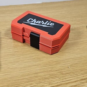 Op de afbeelding: Een rode en zwarte plastic gereedschapskist met de naam "Charlie" op de deksel. De kist is gesloten en staat op een houten oppervlak.
