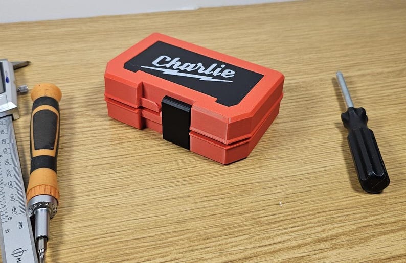 Op de afbeelding: Een rode en zwarte plastic gereedschapskist met de naam "Charlie" op de deksel. De kist is gesloten en staat op een houten oppervlak.