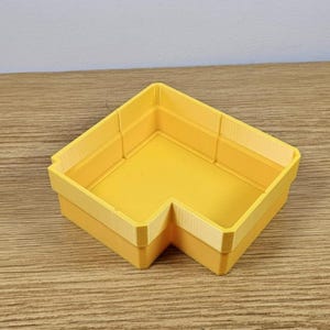 May include: A bright yellow, L-shaped storage container. The container has a smooth, flat interior and a slightly textured exterior. The walls are a lighter shade of yellow, creating a subtle contrast. The container is designed to fit neatly into a corner.