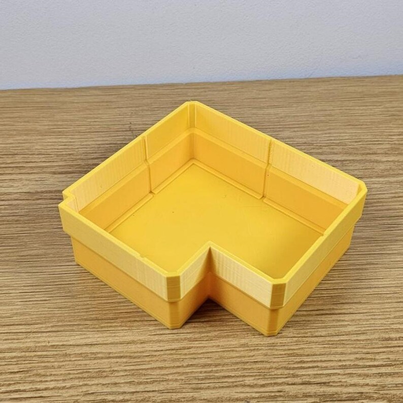 May include: A bright yellow, L-shaped storage container. The container has a smooth, flat interior and a slightly textured exterior. The walls are a lighter shade of yellow, creating a subtle contrast. The container is designed to fit neatly into a corner.