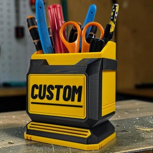Puede incluir: Un organizador de escritorio de plástico amarillo y negro con forma rectangular y diseño escalonado. La parte frontal presenta la palabra CUSTOM en letras negras en negrita. El organizador contiene varias herramientas, incluyendo tijeras naranjas.