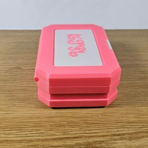 May include: A pink plastic case with the word "Aegis" printed on the top. The case is closed and has a white interior.