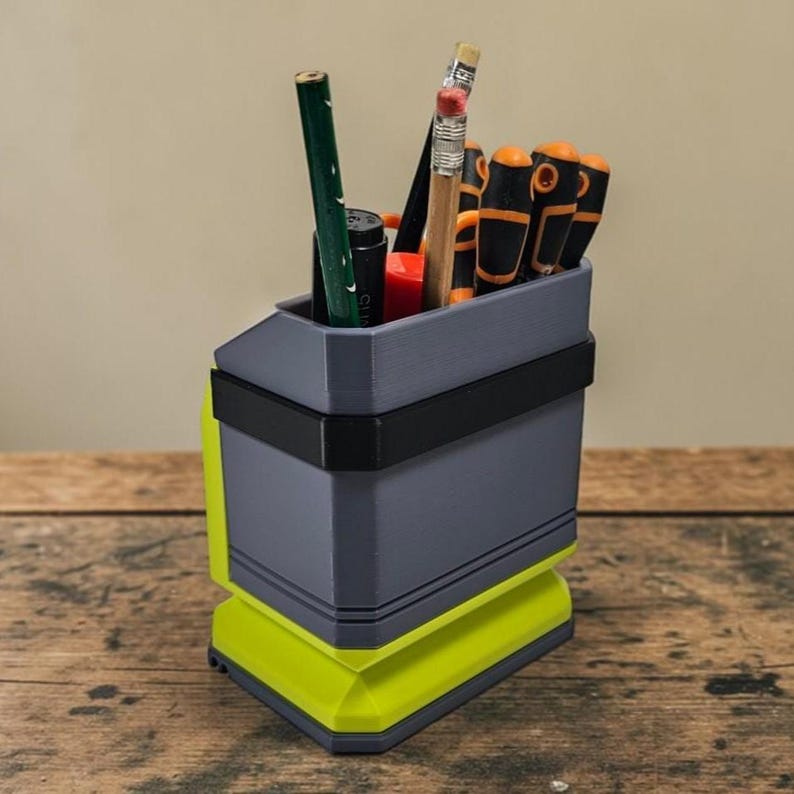 May include: A grey and neon green desk tidy containing pencils, pens, and small tools. The organiser has a geometric design with a black band around the middle. The base is neon green, and the top section is grey.