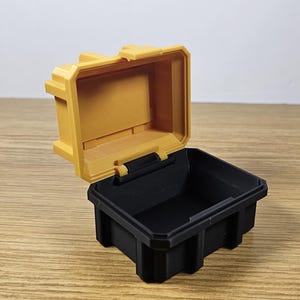 May include: A small, yellow plastic storage box with a black lid. The lid is open and the box is empty.