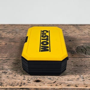 May include: A yellow and black rectangular box with the word "CUSTOM" printed in black. The box has a hinged lid and a black base. The box is sitting on a wooden surface.
