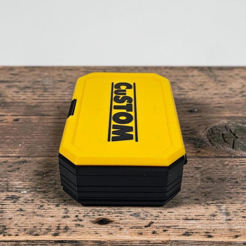 May include: A yellow and black rectangular box with the word "CUSTOM" printed in black. The box has a hinged lid and a black base. The box is sitting on a wooden surface.
