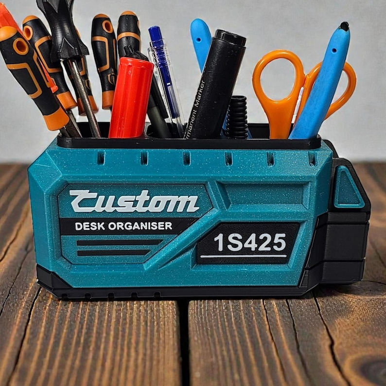 May include: A teal desk organizer with the word "Custom" and "DESK ORGANISER" printed on the front. The organizer holds various tools, including screwdrivers, pens, scissors, and markers. The number "1S425" is printed on the front.