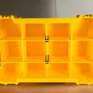 May include: A bright yellow plastic storage organizer insert featuring a grid of nine rectangular compartments. The interior structure includes vertical and horizontal dividers with small black clips securing the center sections, and the DeWalt logo is embossed.