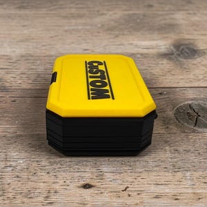 May include: A yellow and black rectangular box with the word "CUSTOM" printed in black on the top. The box has a black base and a yellow lid. The box is sitting on a wooden surface.
