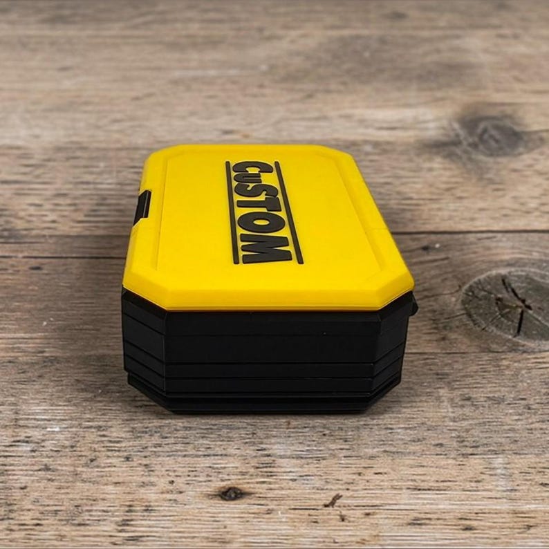 May include: A yellow and black rectangular box with the word "CUSTOM" printed in black on the top. The box has a black base and a yellow lid. The box is sitting on a wooden surface.