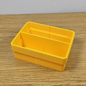 May include: A bright yellow, rectangular organizer with two compartments. The organizer is made of plastic and has a simple, functional design. Ideal for storing small items, it sits on a wooden surface.