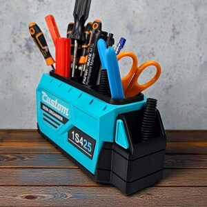 May include: A teal and black desk organizer filled with tools and writing utensils. The organizer has the text "Custom DESK ORGANISER" and the number "1S425" printed on it. Tools include screwdrivers, scissors, and markers.