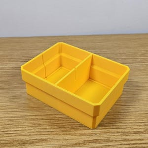 May include: A bright yellow, rectangular storage container with two internal dividers. The container is made of a solid material and has a clean, modern design. The container is sitting on a wooden surface.
