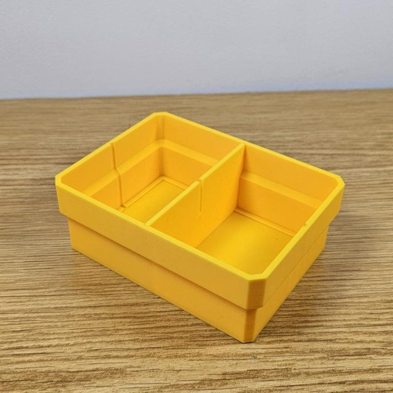 May include: A bright yellow, rectangular storage container with two internal dividers. The container is made of a solid material and has a clean, modern design. The container is sitting on a wooden surface.