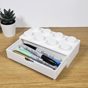 May include: White plastic organizer shaped like a Lego brick with a drawer. The drawer is open and contains four markers, including a black Sharpie Fine Point marker.