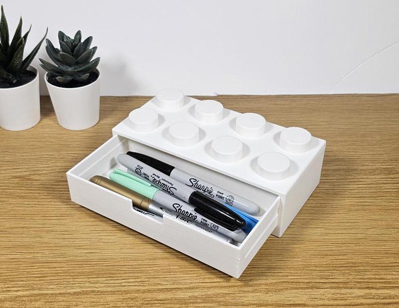 May include: White plastic organizer shaped like a Lego brick with a drawer. The drawer is open and contains four markers, including a black Sharpie Fine Point marker.