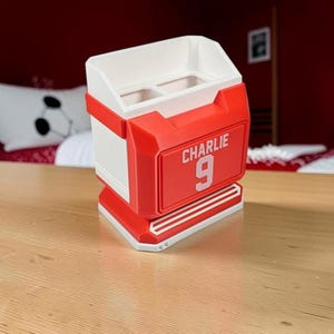 May include: A white and red desk organizer shaped like a sports jersey. The front features a raised red rectangular panel with the white text CHARLIE and the number 9 printed in a bold, athletic font. The top has two open compartments for storage.