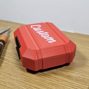 May include: A red plastic case with the word "CUSTOM" printed in white on the top. The case is closed and has a black latch on the side.