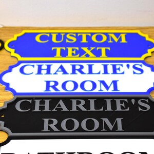 Puede incluir: Primer plano de varios letreros decorativos para habitaciones. El letrero superior es azul con texto amarillo que dice "CUSTOM TEXT". Debajo hay un letrero blanco con texto azul que dice "CHARLIE'S ROOM". Un letrero negro con texto gris tambi&eacute;n dice "CHARLIE'S ROOM".
