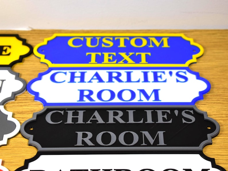 Puede incluir: Primer plano de varios letreros decorativos para habitaciones. El letrero superior es azul con texto amarillo que dice "CUSTOM TEXT". Debajo hay un letrero blanco con texto azul que dice "CHARLIE'S ROOM". Un letrero negro con texto gris tambi&eacute;n dice "CHARLIE'S ROOM".