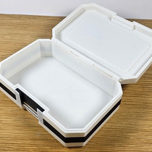 Puede incluir: Caja de pl&aacute;stico blanca con ribete negro. La caja est&aacute; abierta y el interior est&aacute; vac&iacute;o.