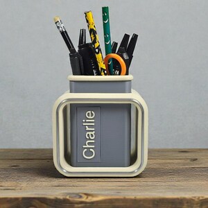May include: A personalized desk organizer with the name "Charlie" printed on the front. The organizer is a square shape with a gray interior and a cream-colored frame. It holds pens, pencils, and scissors.