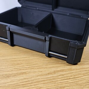 May include: A black plastic storage box with two compartments and a silver metal latch. The box is open and the inside is empty.