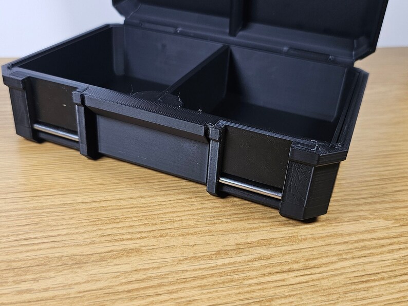 May include: A black plastic storage box with two compartments and a silver metal latch. The box is open and the inside is empty.