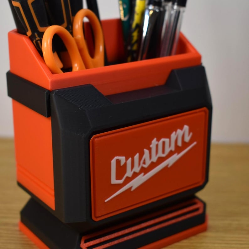 May include: An orange and black desk organizer with the word "Custom" in white script. The organizer holds pens, markers, and orange scissors. The design is reminiscent of a toolbox.
