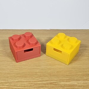 May include: Two small storage boxes shaped like Lego bricks. The box on the left is red and the box on the right is yellow.