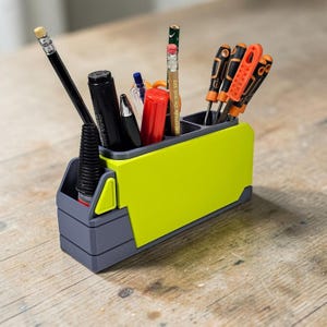 May include: A two-tone desk organizer in gray and lime green, holding various writing utensils and small tools. The organizer has multiple compartments and is made of plastic. A pencil, pens, and screwdrivers are visible.