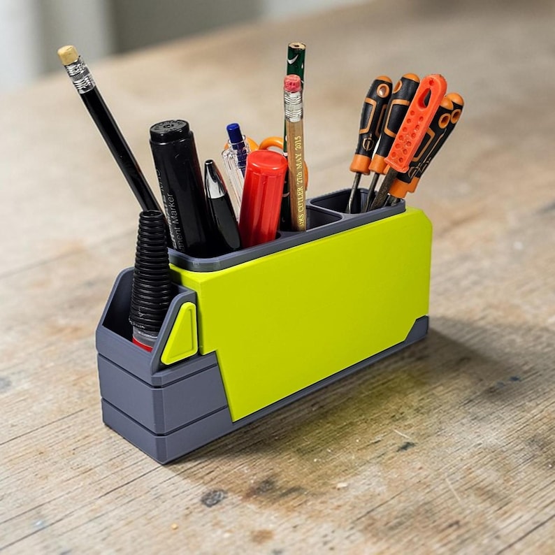 May include: A two-tone desk organizer in gray and lime green, holding various writing utensils and small tools. The organizer has multiple compartments and is made of plastic. A pencil, pens, and screwdrivers are visible.