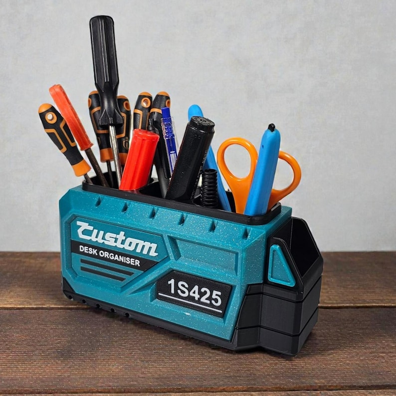 May include: A teal desk organizer with the word "Custom" and "DESK ORGANISER" printed on the front. The organizer holds various tools, including screwdrivers, pens, and scissors. The number "1S425" is printed on the front.
