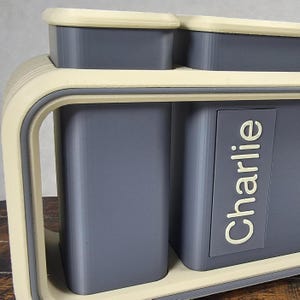 May include: A two-compartment storage container with a light beige frame and trim, and gray interior compartments. The word "Charlie" is printed in white on the side of one compartment. The container has a modern, minimalist design.