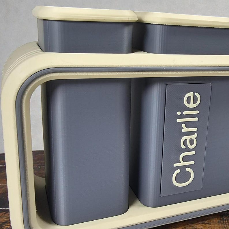 May include: A two-compartment storage container with a light beige frame and trim, and gray interior compartments. The word "Charlie" is printed in white on the side of one compartment. The container has a modern, minimalist design.