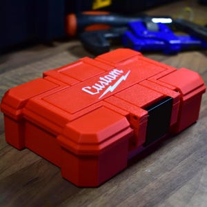 May include: A red tool case with the word "Custom" in white script and a lightning bolt logo. The case has a rectangular shape with a black latch and is sitting on a wooden surface. Other tools are visible in the background.