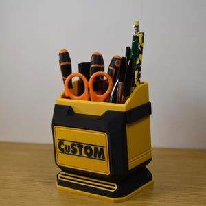 May include: A yellow and black desk organizer with the word "CUSTOM" printed on the front. The organizer holds various tools, including screwdrivers, scissors, pens, and pencils. The organizer is made of plastic and sits on a wooden surface.