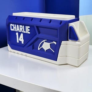 May include: A rectangular blue and white desk organizer featuring the name CHARLIE and the number 14 in white lettering. The front panel displays a white graphic of a soccer ball. The structure has multiple compartments for storage.