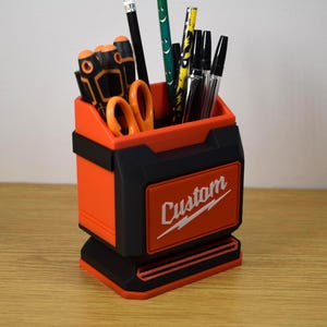 May include: An orange and black desk organizer with the word "Custom" in white script. The organizer holds pencils, pens, and scissors. The design is reminiscent of a toolbox.