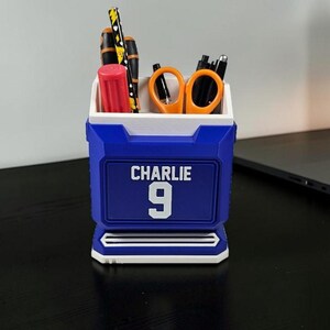 May include: A blue and white desk organizer with the name "Charlie" and the number "9" on the front. The organizer holds pens, scissors, and a red cylinder. The design is reminiscent of a sports jersey.
