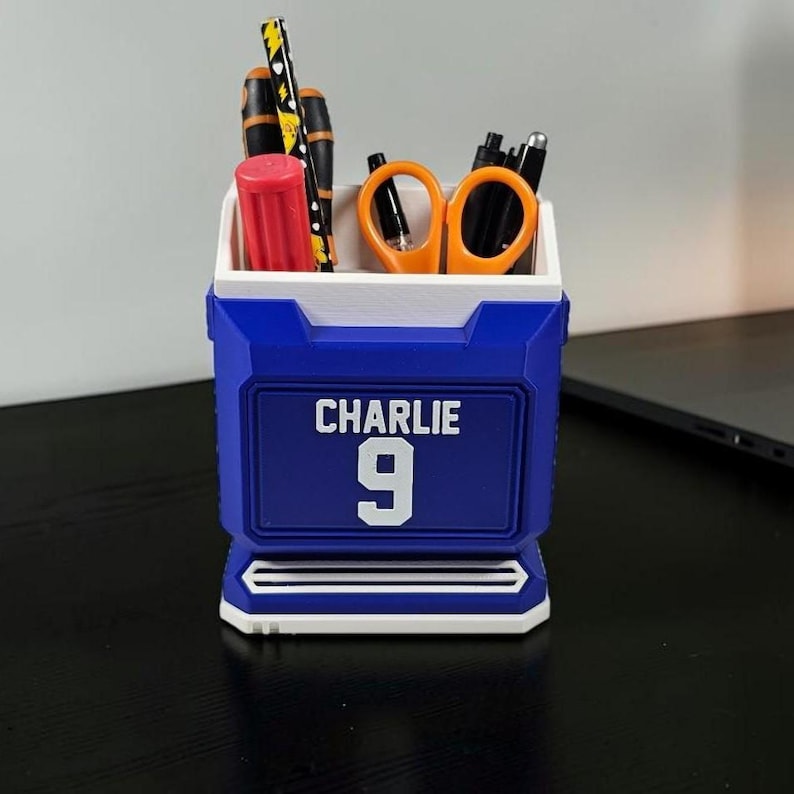 May include: A blue and white desk organizer with the name "Charlie" and the number "9" on the front. The organizer holds pens, scissors, and a red cylinder. The design is reminiscent of a sports jersey.