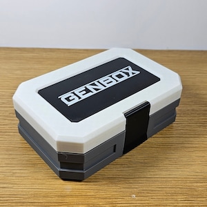 K&ouml;nnte beinhalten: Eine wei&szlig;e und graue Plastik-Aufbewahrungsbox mit einem schwarzen Verschluss und dem Wort "GENBOX" auf der Oberseite.