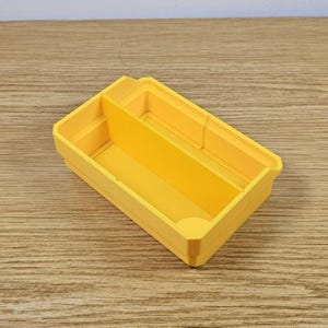 May include: A bright yellow rectangular organizer with two compartments. The organizer is made of plastic and sits on a wooden surface. The compartments are divided by a vertical divider, creating two separate storage spaces.