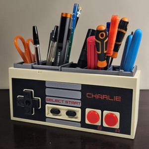 May include: A beige and black desk organizer shaped like a classic video game controller, with the name "CHARLIE" printed on it. The organizer holds pens, scissors, and other office supplies. The controller design includes buttons and directional pad.