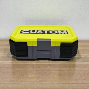 May include: A rectangular storage box with a yellow lid and black and gray body. The word "CUSTOM" is printed in bold white letters on the lid. The box is sitting on a wooden surface.