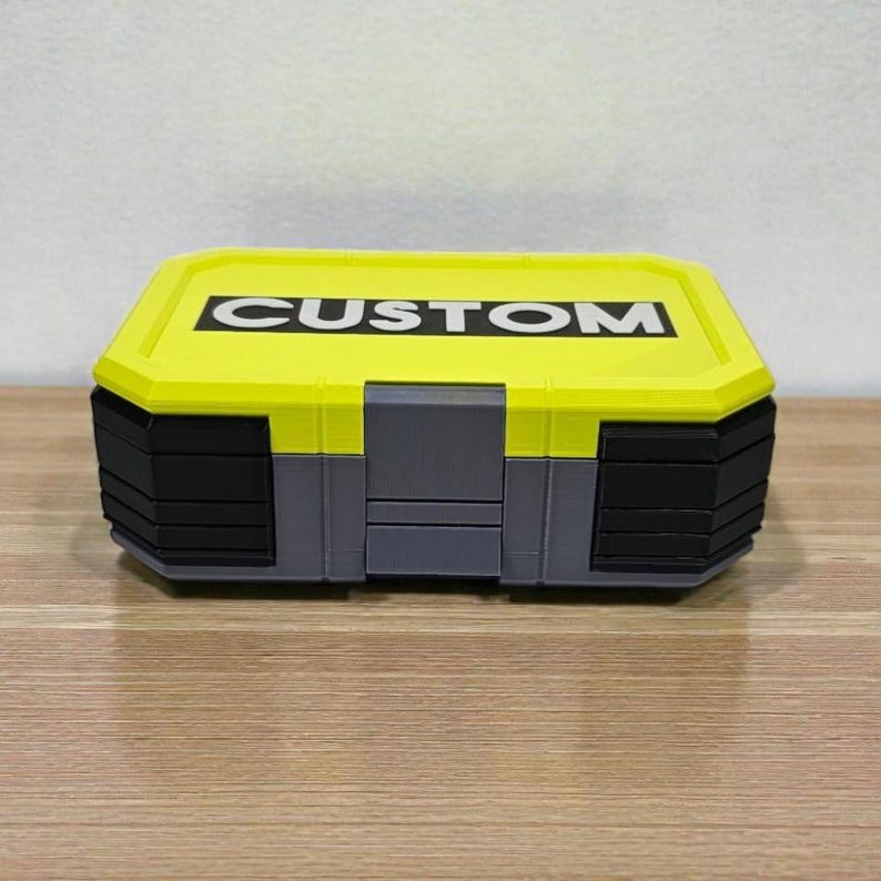 May include: A rectangular storage box with a yellow lid and black and gray body. The word "CUSTOM" is printed in bold white letters on the lid. The box is sitting on a wooden surface.