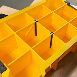 May include: A bright yellow plastic storage organizer box featuring multiple rectangular compartments separated by removable dividers. The container includes black plastic locking latches on the sides and a grid-like interior structure for sorting small items.
