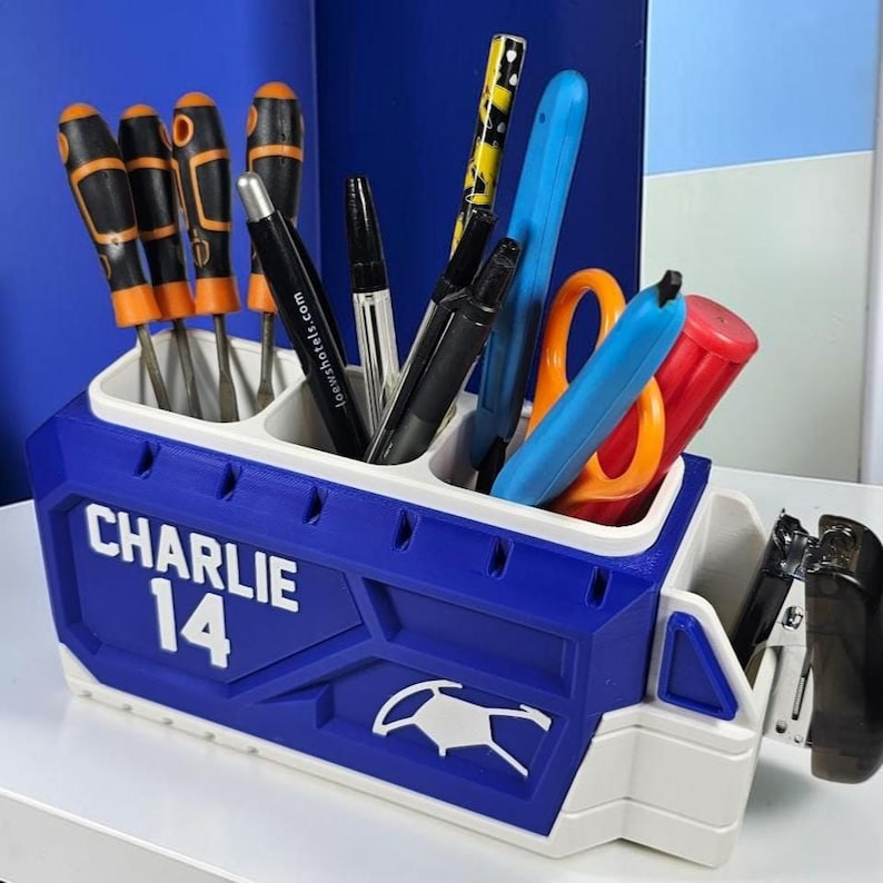 May include: A blue and white desk organizer shaped like a truck, featuring the text CHARLIE 14 on the side. The organizer has multiple compartments filled with various office supplies, including screwdrivers, pens, scissors, and a stapler.