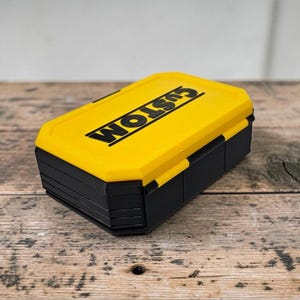 May include: A yellow and black storage box with the word "CUSTOM" printed on the lid. The box is rectangular with a hinged lid and a black base. The box is sitting on a wooden surface.
