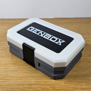 K&ouml;nnte beinhalten: Eine wei&szlig;e und graue Aufbewahrungsbox mit einem schwarzen Deckel. Der Deckel tr&auml;gt den Schriftzug "GENBOX" in wei&szlig;en Buchstaben.